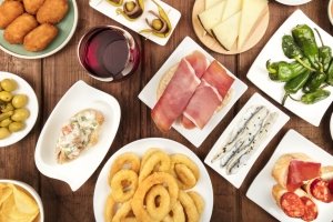 The food of Spain. An overhead photo of many different Spanish tapas, shot from above on a dark rustic texture. Jamon, cheese, wine, olives, croquettes, calamari rings, anchovies and more. Concept of Wine and Tapas pairing.