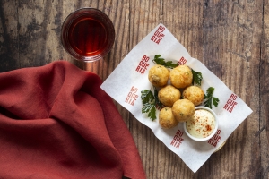 Salt cod and potato fritters on a white sheet of parchment paper served with a garnish of parsley, citrus aioli, and a beer.